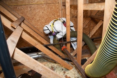 A worker wearing a white tyvek protective suit decontaminates an area in the attic of a home.